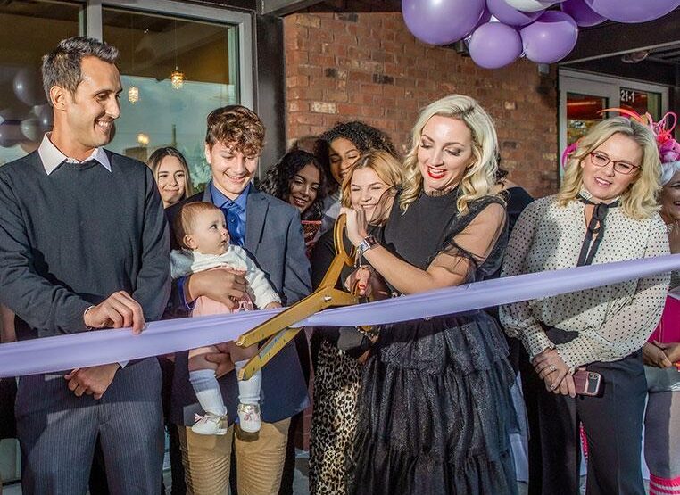 A group of lash business owners cutting a purple ribbon in front of balloons.
